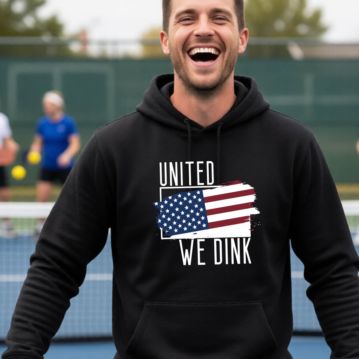 A man on a pickleball court wearing a black hoodie pullover with a pickleball themed red, white and blue graphic of a patriotic flag with white words “UNITED WE DINK” above and below the flag, on the front of the hoodie.