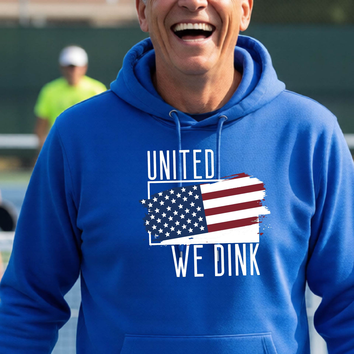 A man on a pickleball court wearing a royal blue  hoodie pullover with a pickleball themed red, white and blue graphic of a patriotic flag with white words “UNITED WE DINK” above and below the flag, on the front of the hoodie.