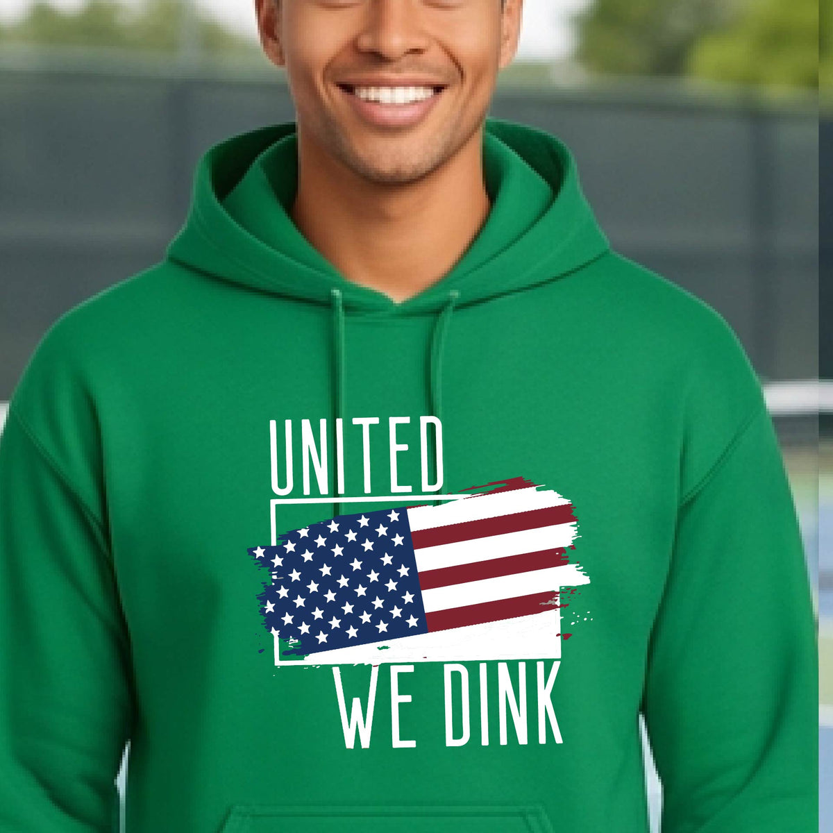 A man on a pickleball court wearing a green hoodie pullover with a pickleball themed red, white and blue graphic of a patriotic flag with white words “UNITED WE DINK” above and below the flag, on the front of the hoodie.
