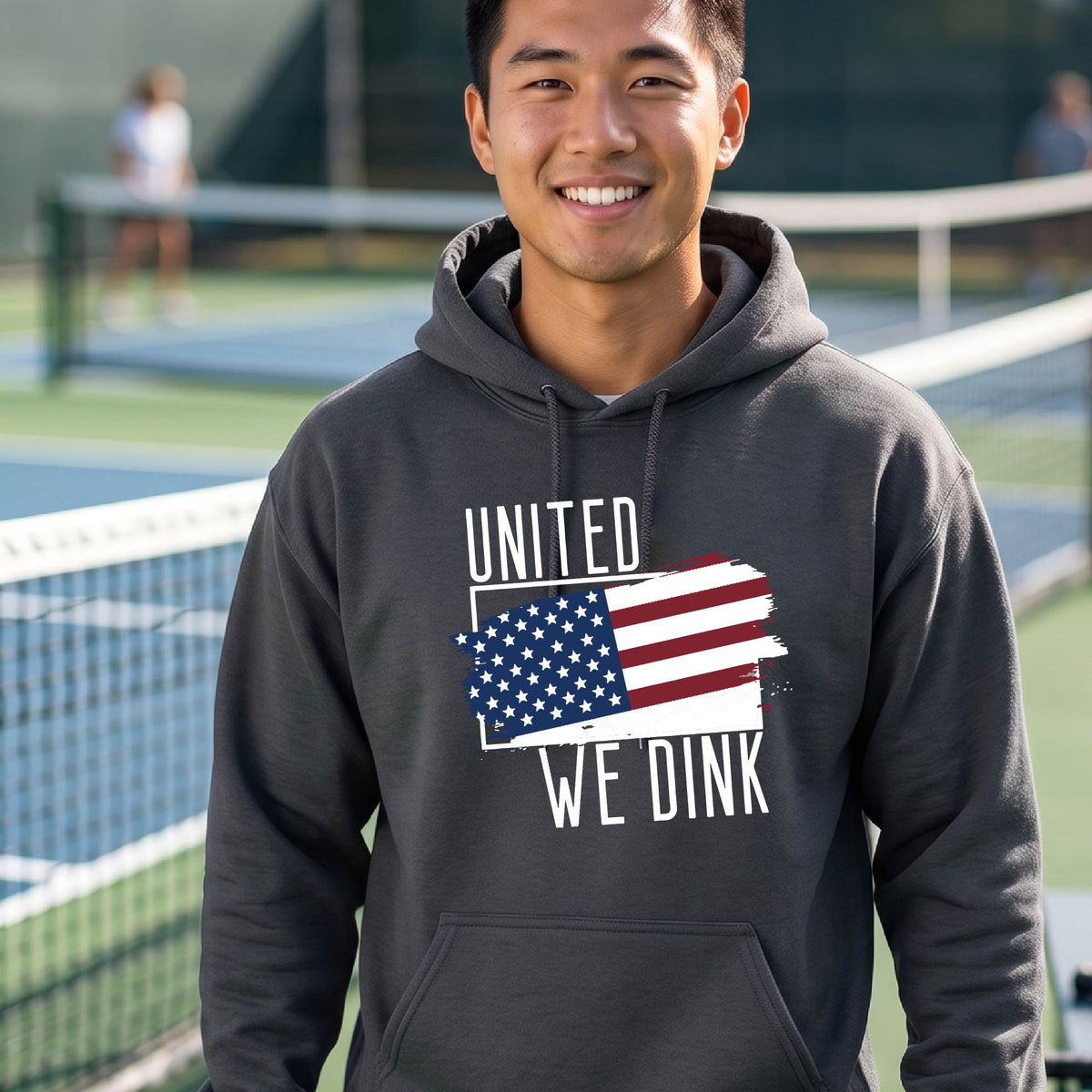 A man on a pickleball court wearing a gray hoodie pullover with a pickleball themed red, white and blue graphic of a patriotic flag with white words “UNITED WE DINK” above and below the flag, on the front of the hoodie.