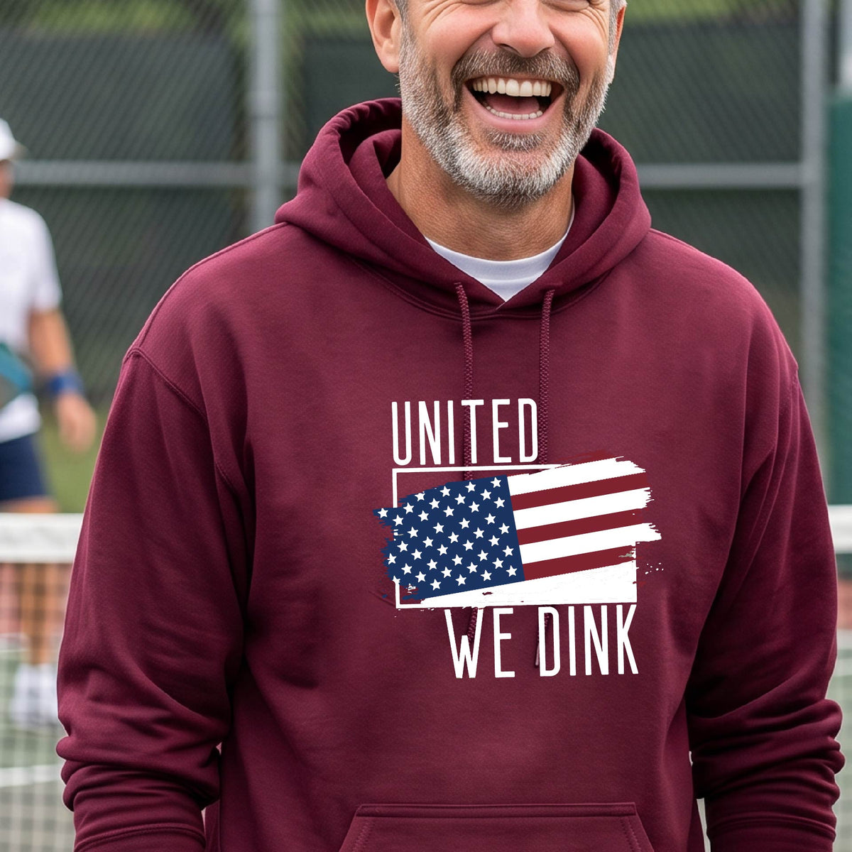 A man on a pickleball court wearing a maroon hoodie pullover with a pickleball themed red, white and blue graphic of a patriotic flag with white words “UNITED WE DINK” above and below the flag, on the front of the hoodie.
