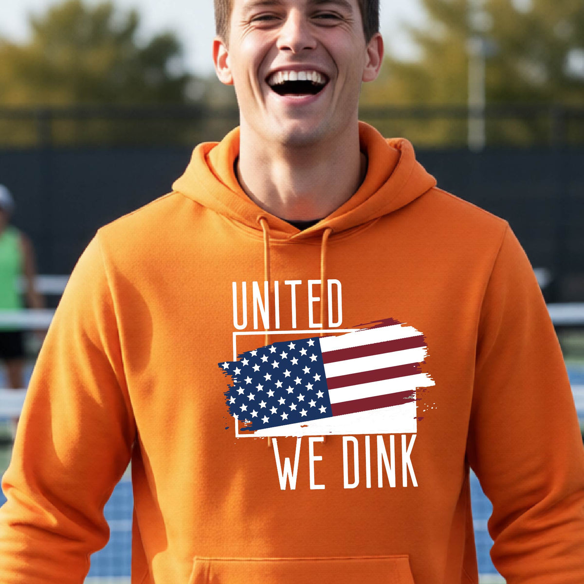 A man on a pickleball court wearing an orange hoodie pullover with a pickleball themed red, white and blue graphic of a patriotic flag with white words “UNITED WE DINK” above and below the flag, on the front of the hoodie.