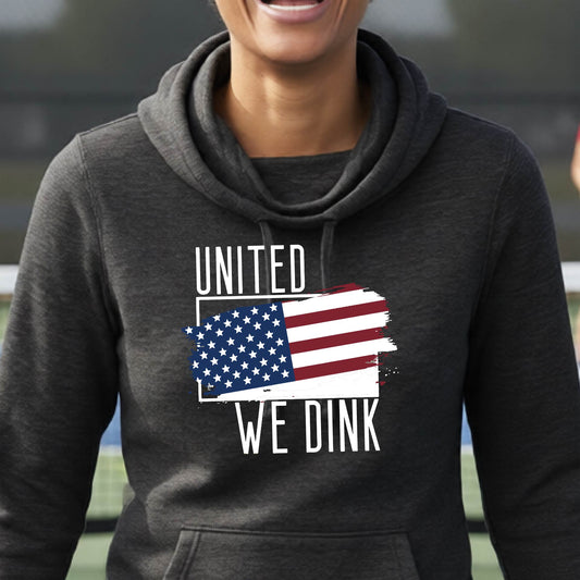 A woman on a pickleball court wearing a black hoodie pullover with a pickleball themed red, white and blue graphic of a patriotic flag with white words “UNITED WE DINK” above and below the flag, on the front of the hoodie.