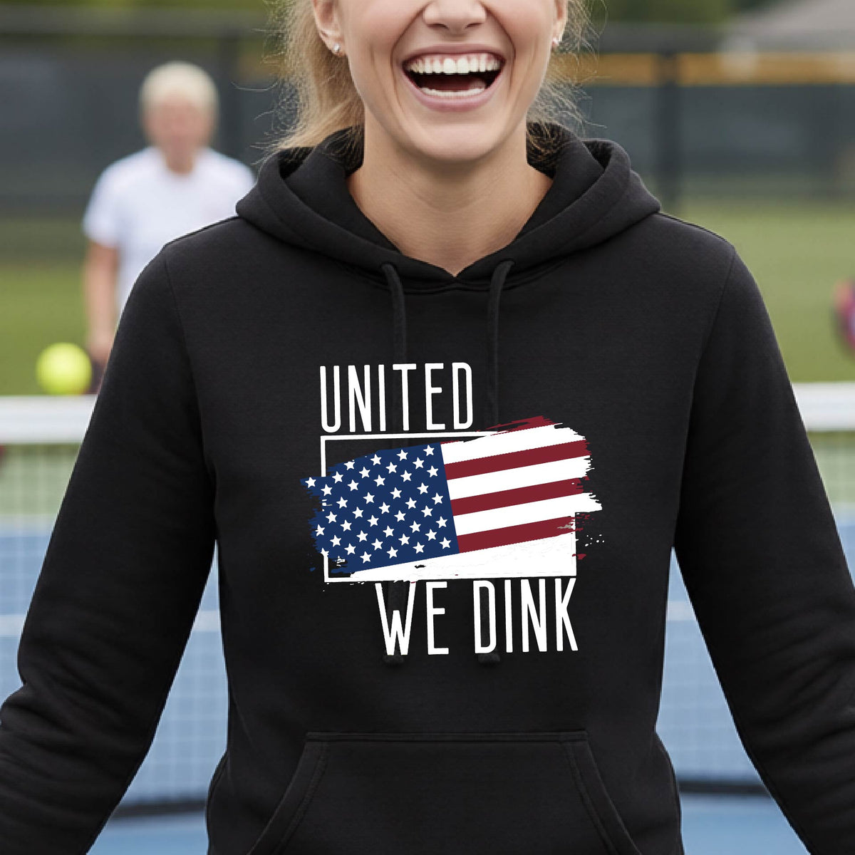A woman on a pickleball court wearing a black hoodie pullover with a pickleball themed red, white and blue graphic of a patriotic flag with white words “UNITED WE DINK” above and below the flag, on the front of the hoodie.
