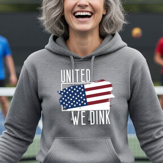 A woman on a pickleball court wearing a gray hoodie pullover with a pickleball themed red, white and blue graphic of a patriotic flag with white words “UNITED WE DINK” above and below the flag, on the front of the hoodie.