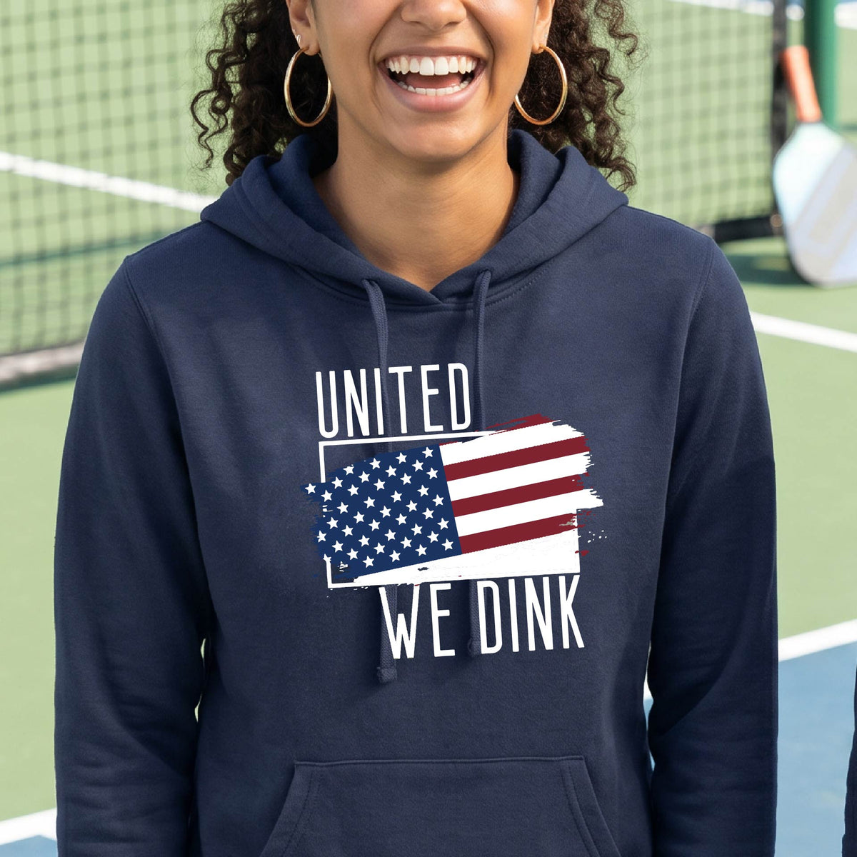 A woman on a pickleball court wearing a navy blue hoodie pullover with a pickleball themed red, white and blue graphic of a patriotic flag with white words “UNITED WE DINK” above and below the flag, on the front of the hoodie.