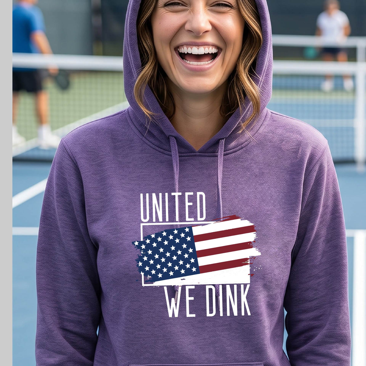 A woman on a pickleball court wearing a purple hoodie pullover with a pickleball themed red, white and blue graphic of a patriotic flag with white words “UNITED WE DINK” above and below the flag, on the front of the hoodie.