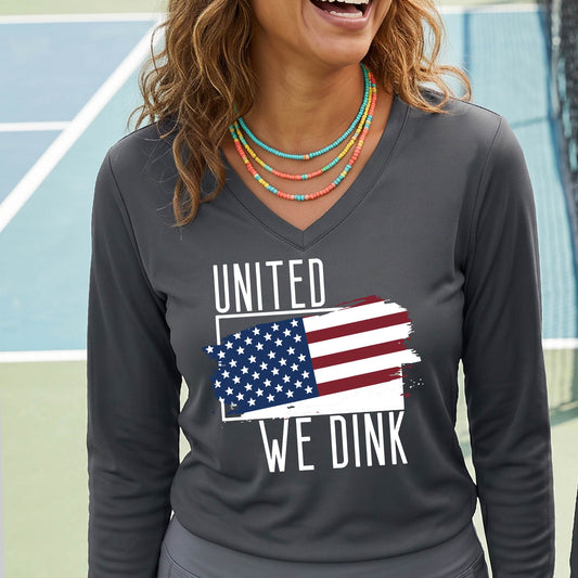 A woman on a pickleball court wearing a gray long-sleeve t-shirt with a pickleball themed red, white and blue graphic of a patriotic flag with white words “UNITED WE DINK” above and below the flag, on the front of the shirt. 