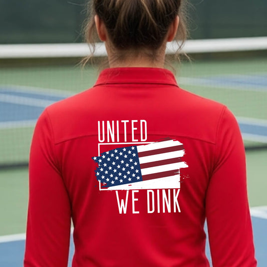 A woman seen from behind on a pickleball court wearing a red quarter-zip shirt with a pickleball themed red, white and blue graphic of a patriotic flag with white words “UNITED WE DINK” above and below the flag on the back of the shirt.