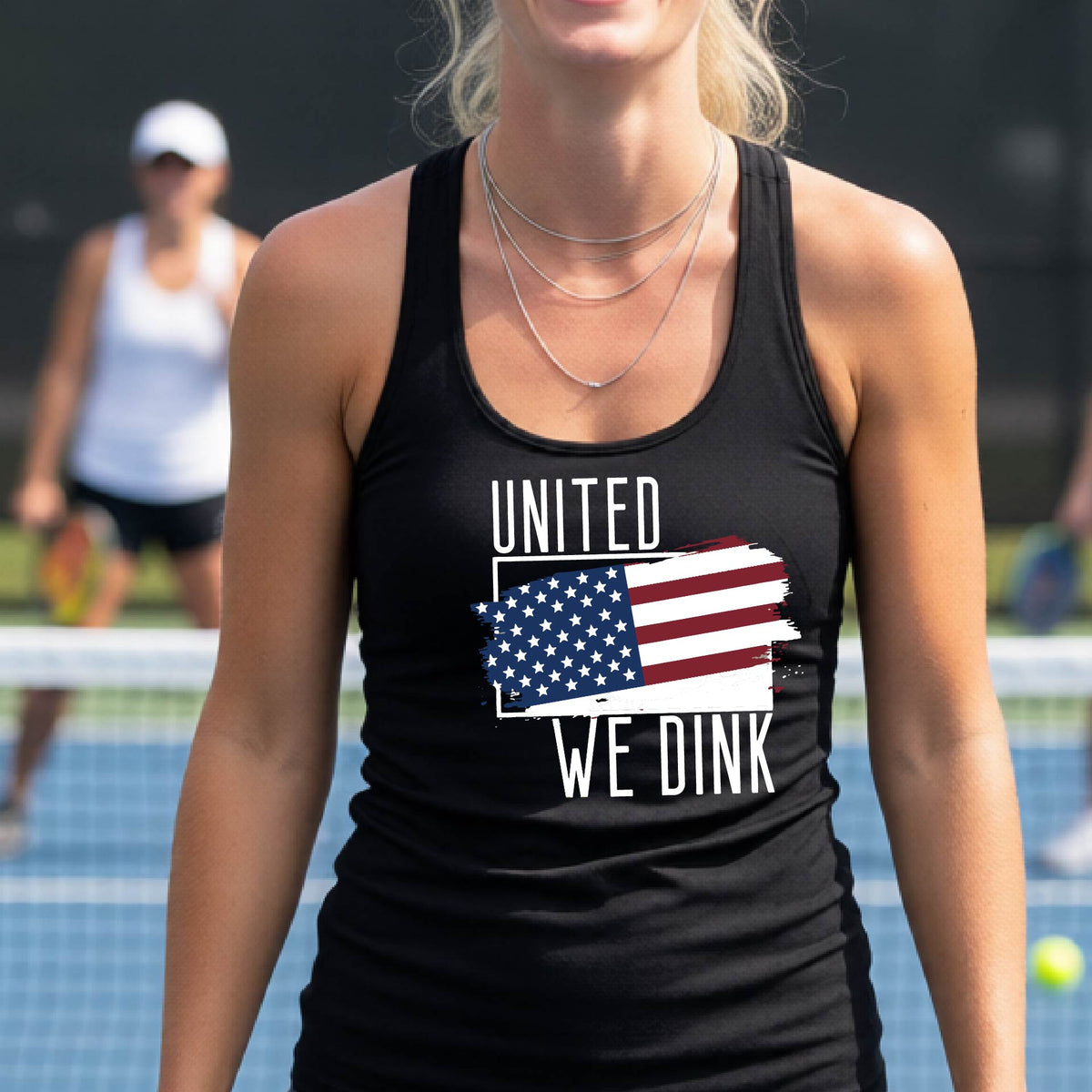 A woman on a pickleball court wearing a black racerback tank top with a pickleball themed red, white and blue graphic of a patriotic flag with white words “UNITED WE DINK” above and below the flag, on the front of the shirt. 