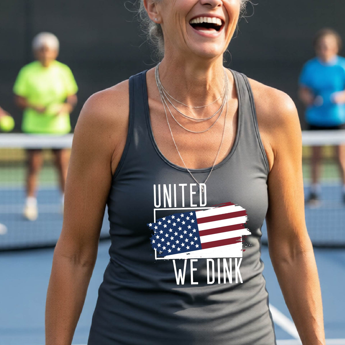 A woman on a pickleball court wearing a gray racerback tank top with a pickleball themed red, white and blue graphic of a patriotic flag with white words “UNITED WE DINK” above and below the flag, on the front of the shirt. 