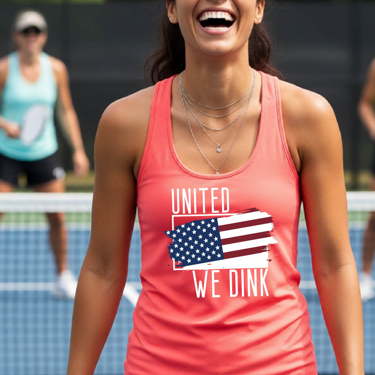 A woman on a pickleball court wearing a coral pink racerback tank top with a pickleball themed red, white and blue graphic of a patriotic flag with white words “UNITED WE DINK” above and below the flag, on the front of the shirt. 