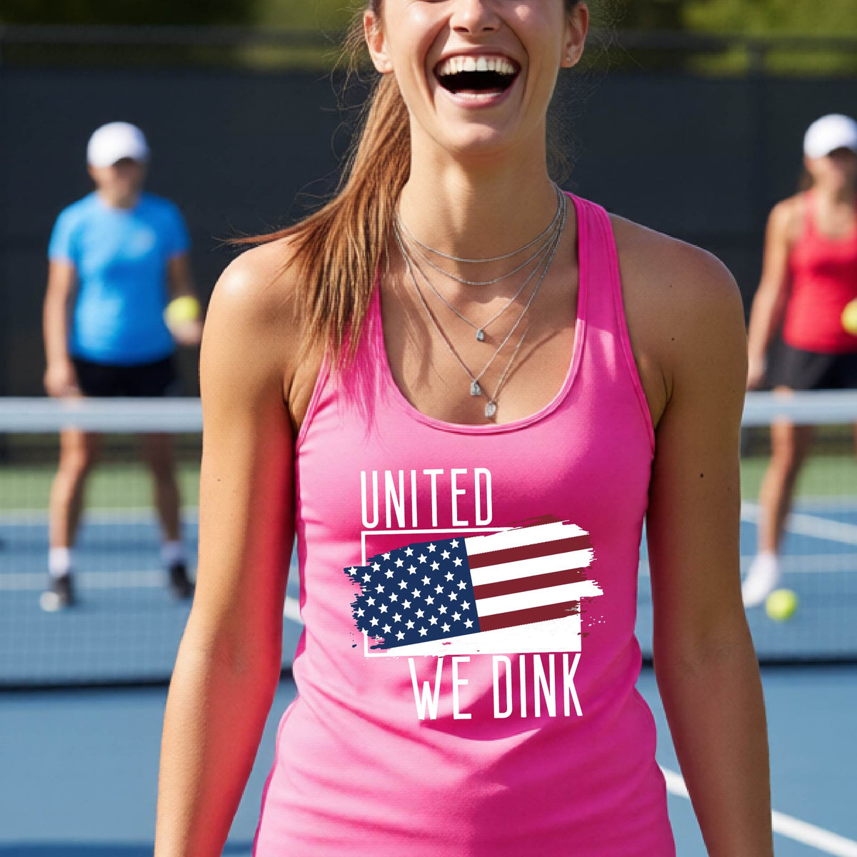 A woman on a pickleball court wearing a pink racerback tank top with a pickleball themed red, white and blue graphic of a patriotic flag with white words “UNITED WE DINK” above and below the flag, on the front of the shirt. 