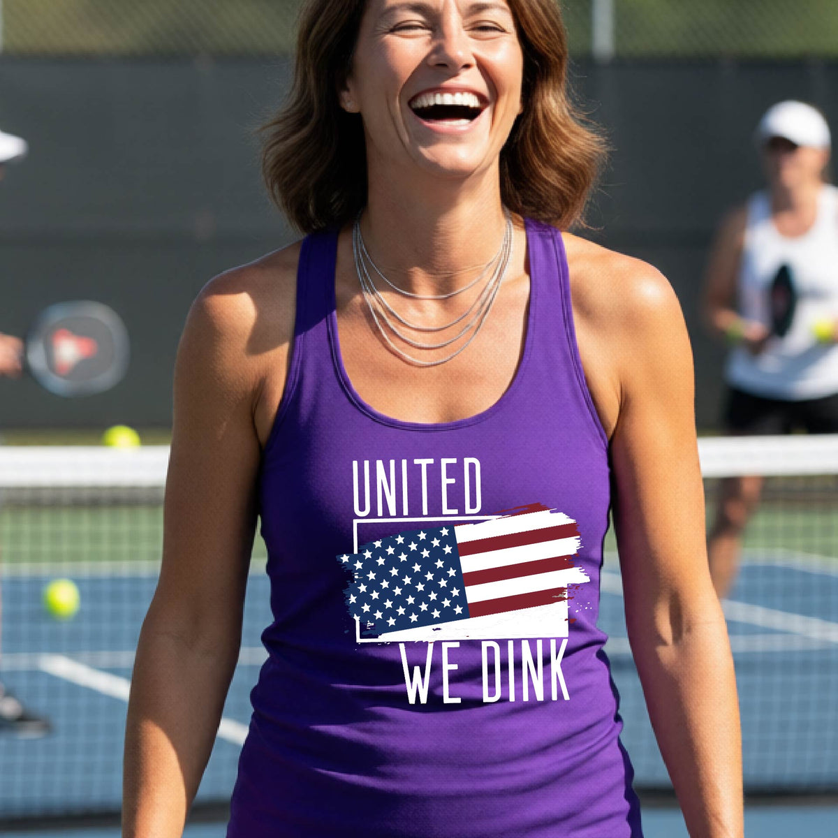 A woman on a pickleball court wearing a purple racerback tank top with a pickleball themed red, white and blue graphic of a patriotic flag with white words “UNITED WE DINK” above and below the flag, on the front of the shirt. 