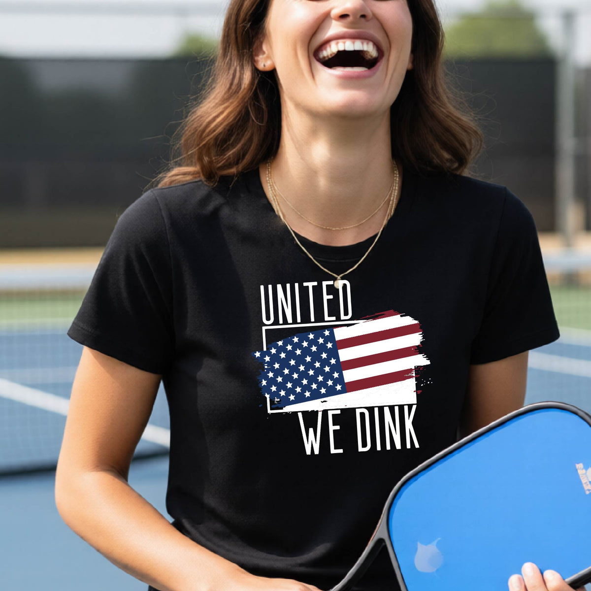 A woman on a pickleball court wearing a black short-sleeve t-shirt with a pickleball themed red, white and blue graphic of a patriotic flag with white words “UNITED WE DINK” above and below the flag, on the front of the shirt. 