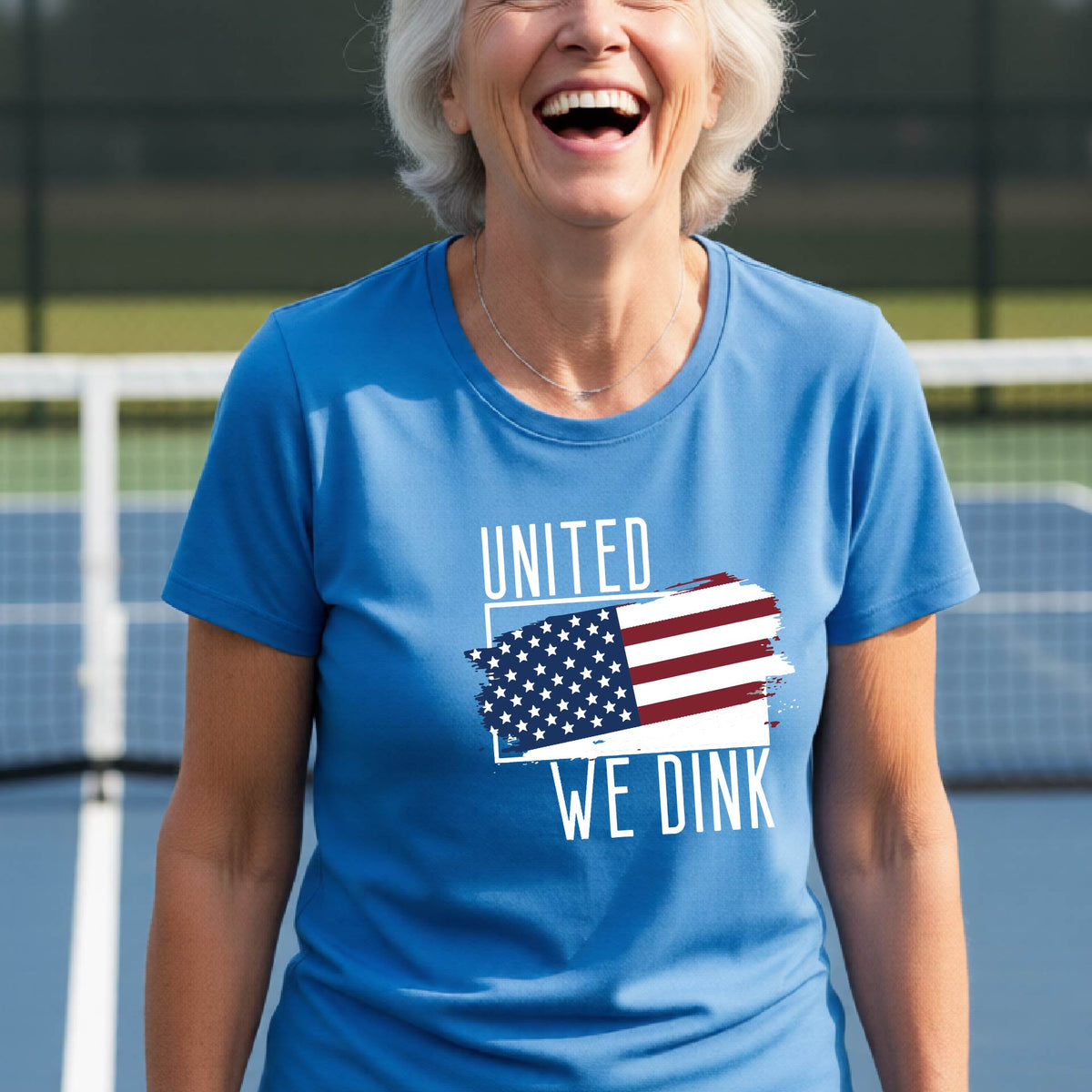 A woman on a pickleball court wearing a blue short-sleeve t-shirt with a pickleball themed red, white and blue graphic of a patriotic flag with white words “UNITED WE DINK” above and below the flag, on the front of the shirt.