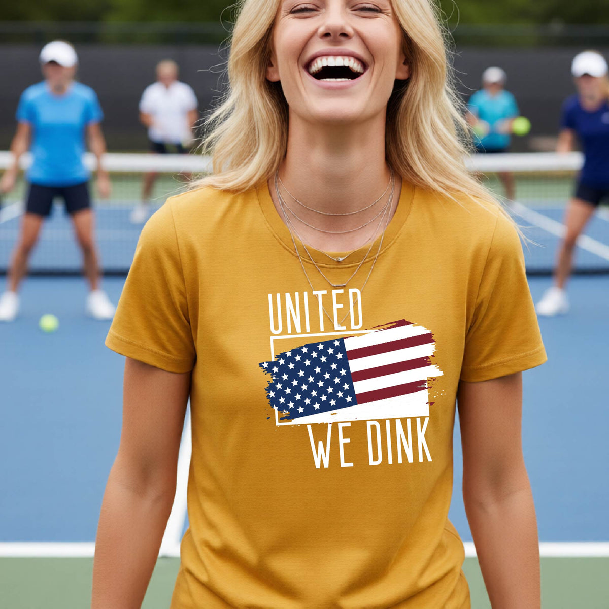A woman on a pickleball court wearing a mustard yellow gold short-sleeve t-shirt with a pickleball themed red, white and blue graphic of a patriotic flag with white words “UNITED WE DINK” above and below the flag, on the front of the shirt. 