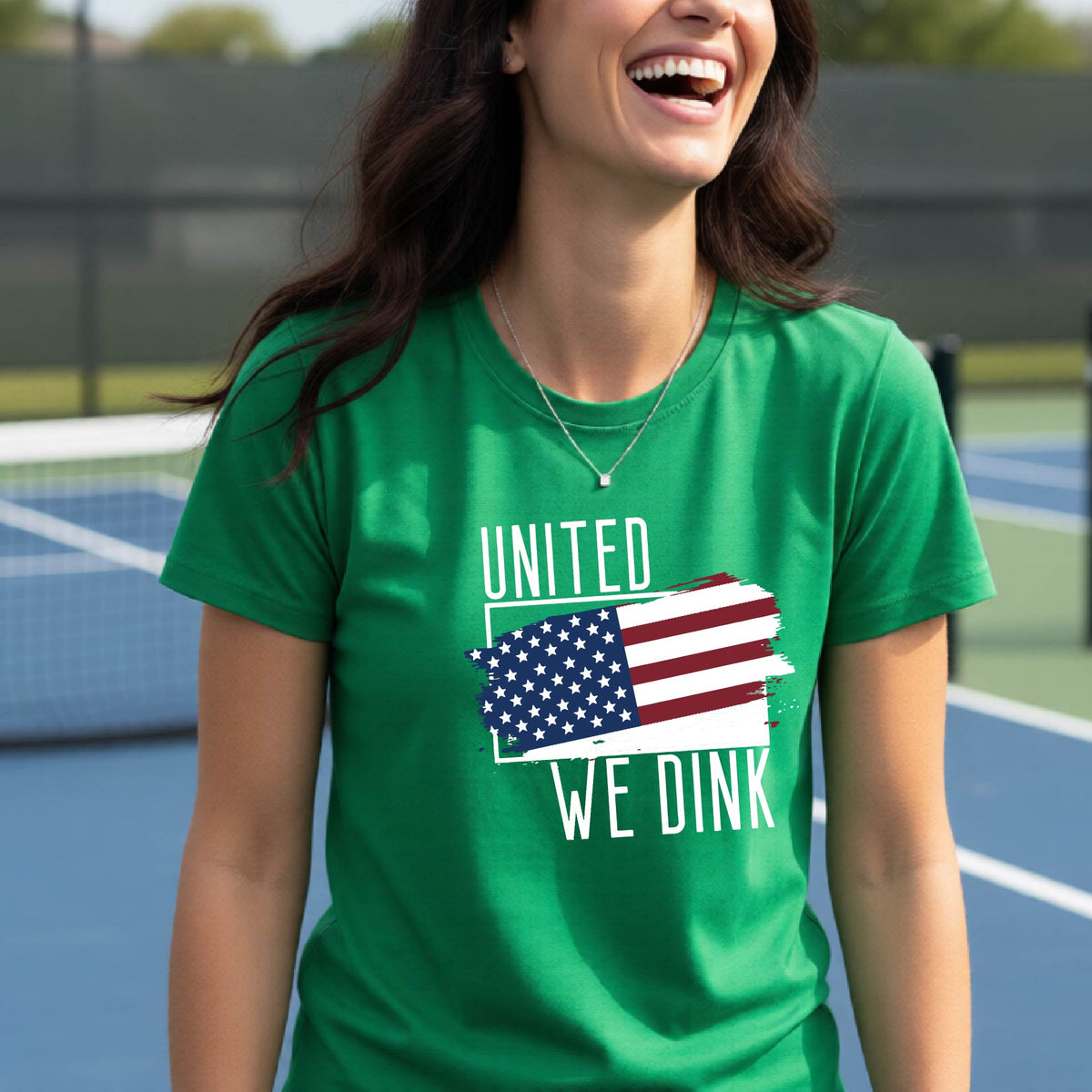 A woman on a pickleball court wearing a green short-sleeve t-shirt with a pickleball themed red, white and blue graphic of a patriotic flag with white words “UNITED WE DINK” above and below the flag, on the front of the shirt.