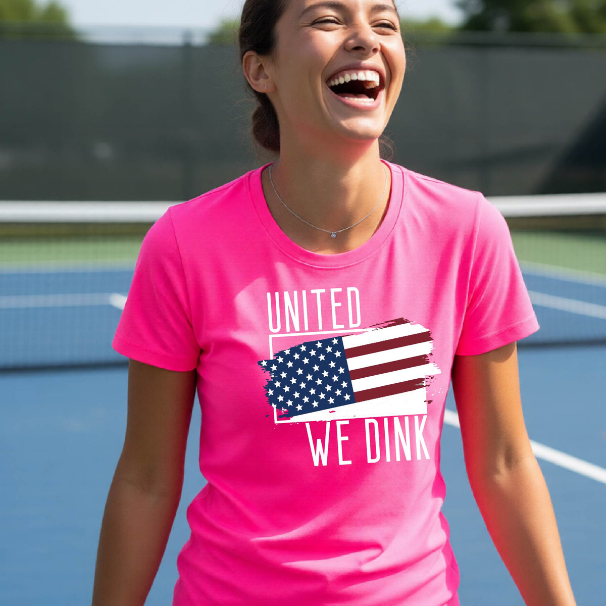 A woman on a pickleball court wearing a pink short-sleeve t-shirt with a pickleball themed red, white and blue graphic of a patriotic flag with white words “UNITED WE DINK” above and below the flag, on the front of the shirt.