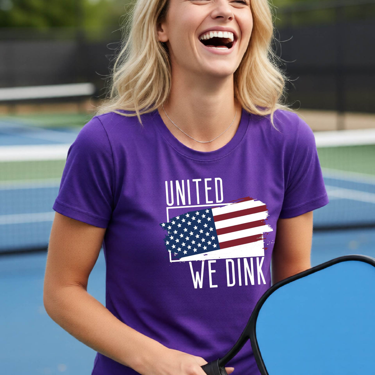 A woman on a pickleball court wearing a purple short-sleeve t-shirt with a pickleball themed red, white and blue graphic of a patriotic flag with white words “UNITED WE DINK” above and below the flag, on the front of the shirt.