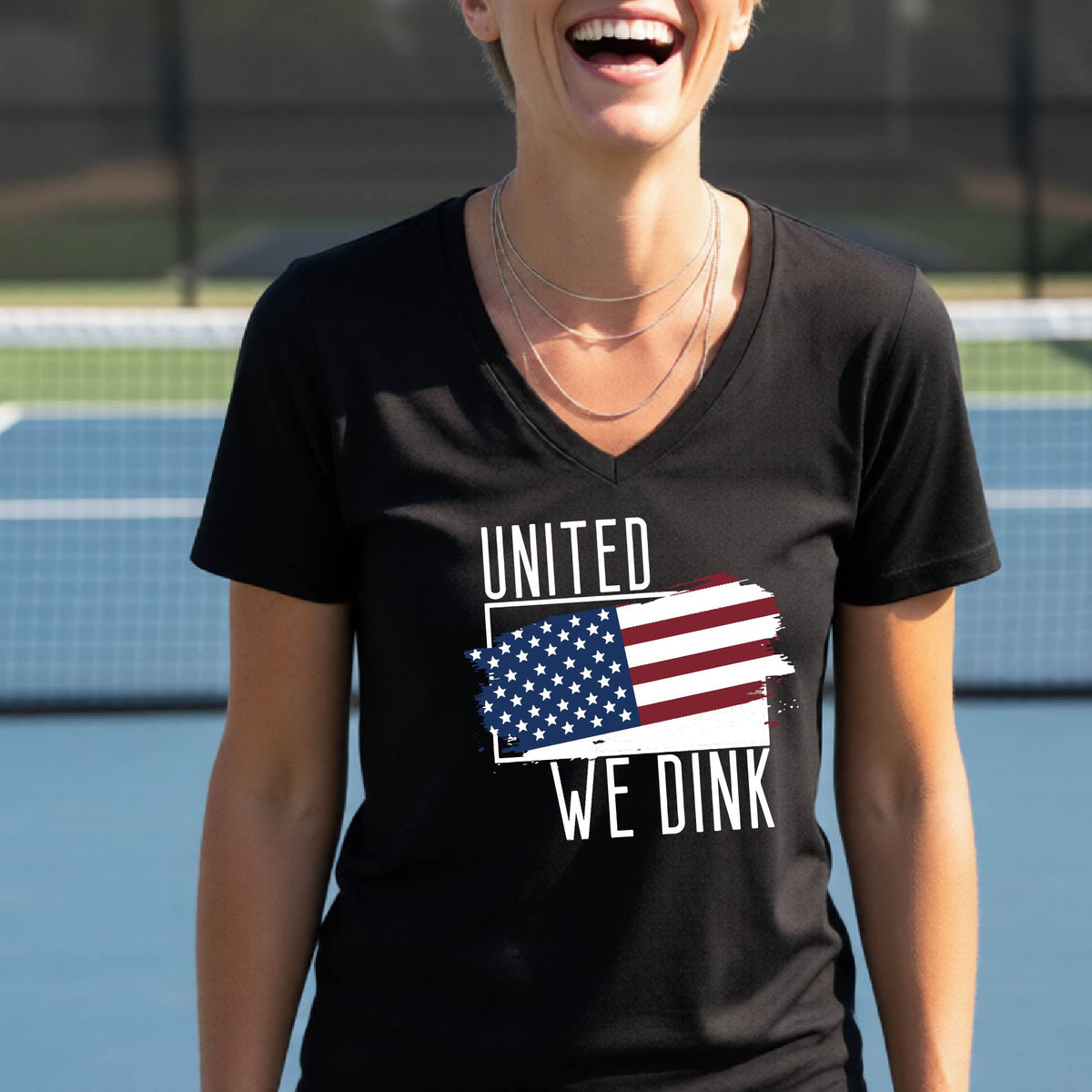 A woman on a pickleball court wearing a black short-sleeve v-neck t-shirt with a pickleball themed red, white and blue graphic of a patriotic flag with white words “UNITED WE DINK” above and below the flag, on the front of the shirt. 