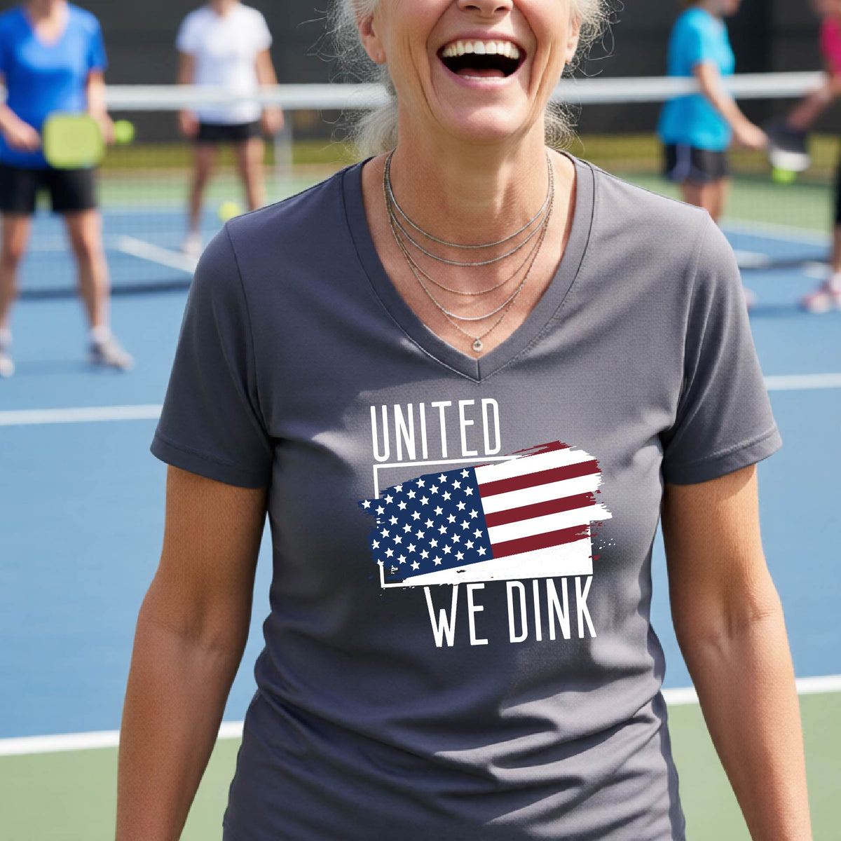 A woman on a pickleball court wearing a gray short-sleeve v-neck t-shirt with a pickleball themed red, white and blue graphic of a patriotic flag with white words “UNITED WE DINK” above and below the flag, on the front of the shirt. 