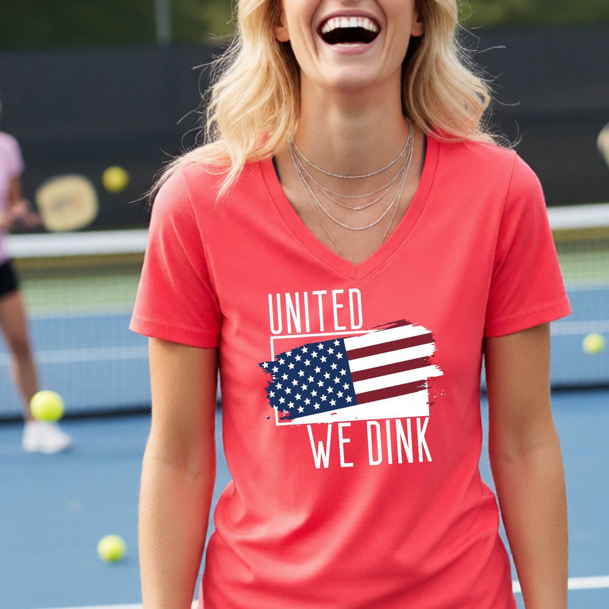 A woman on a pickleball court wearing a coral pink short-sleeve v-neck t-shirt with a pickleball themed red, white and blue graphic of a patriotic flag with white words “UNITED WE DINK” above and below the flag, on the front of the shirt. 
