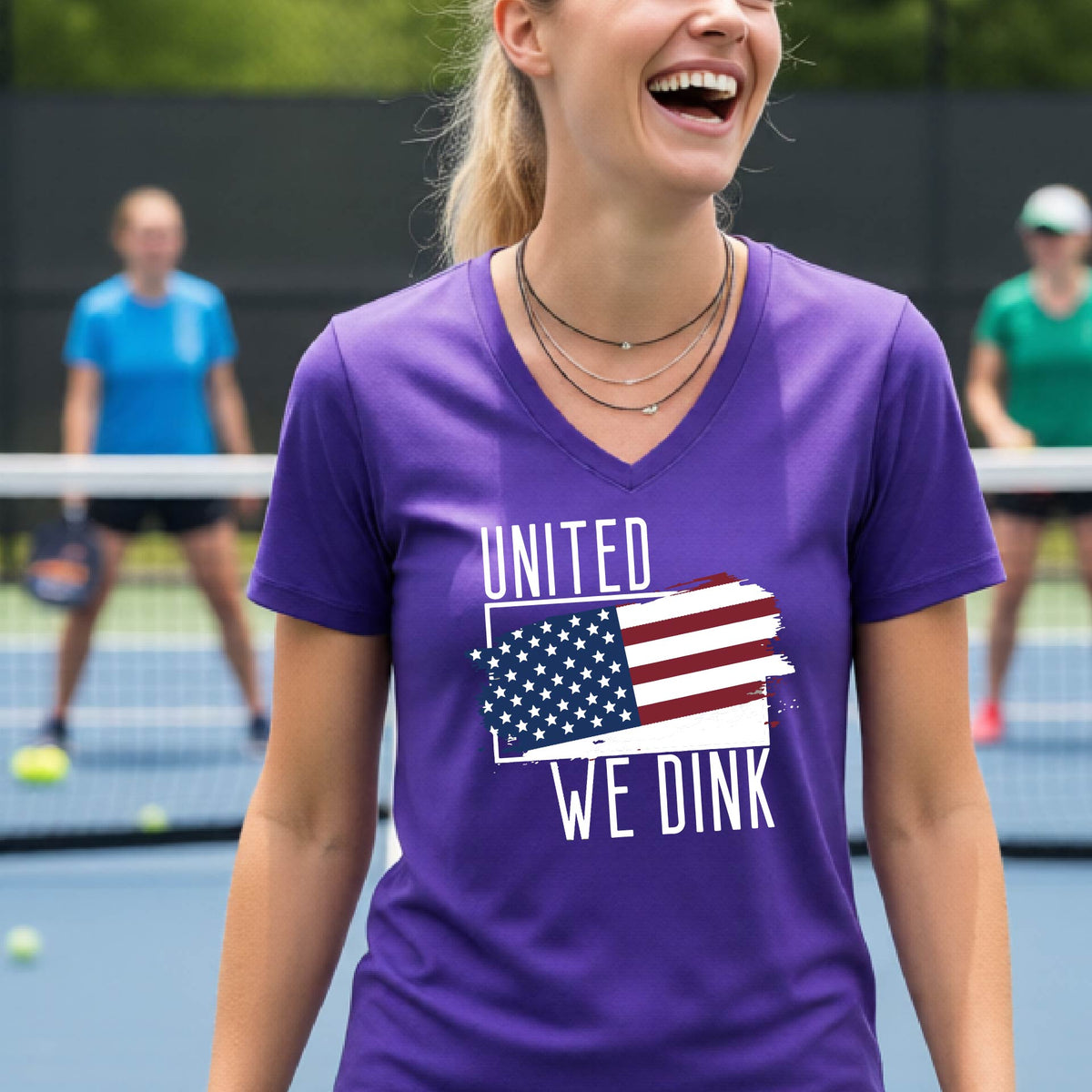 A woman on a pickleball court wearing a purple short-sleeve v-neck t-shirt with a pickleball themed red, white and blue graphic of a patriotic flag with white words “UNITED WE DINK” above and below the flag, on the front of the shirt. 