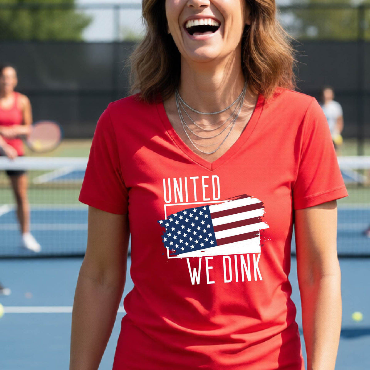 A woman on a pickleball court wearing a red short-sleeve v-neck t-shirt with a pickleball themed red, white and blue graphic of a patriotic flag with white words “UNITED WE DINK” above and below the flag, on the front of the shirt. 