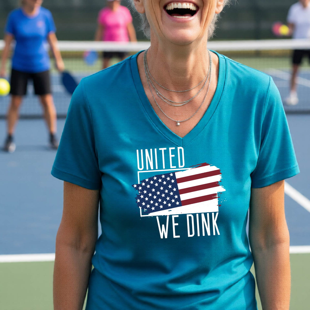 A woman on a pickleball court wearing a teal short-sleeve v-neck t-shirt with a pickleball themed red, white and blue graphic of a patriotic flag with white words “UNITED WE DINK” above and below the flag, on the front of the shirt. 