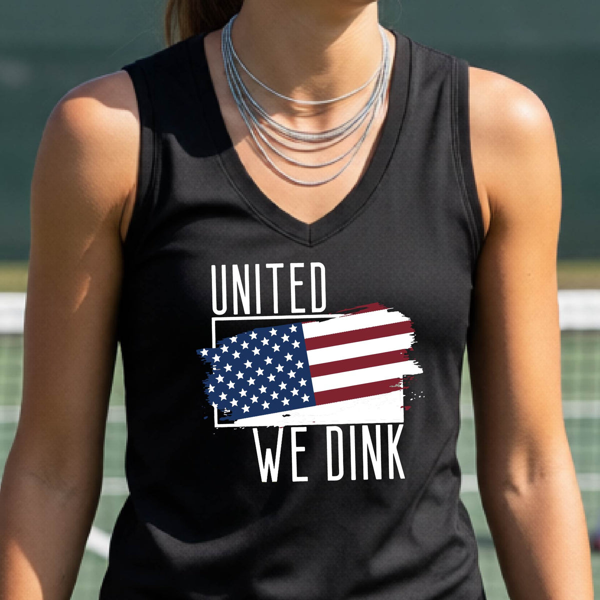 A woman on a pickleball court wearing a black sleeveless t-shirt with a pickleball themed red, white and blue graphic of a patriotic flag with white words “UNITED WE DINK” above and below the flag, on the front of the shirt. 