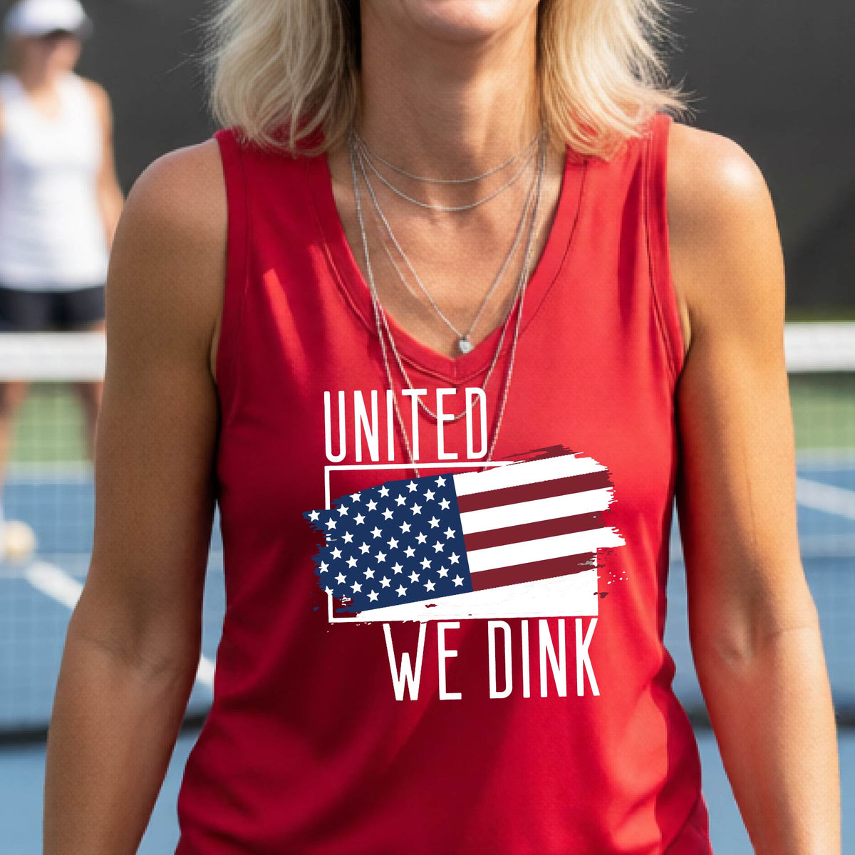 A woman on a pickleball court wearing a red sleeveless t-shirt with a pickleball themed red, white and blue graphic of a patriotic flag with white words “UNITED WE DINK” above and below the flag, on the front of the shirt. 