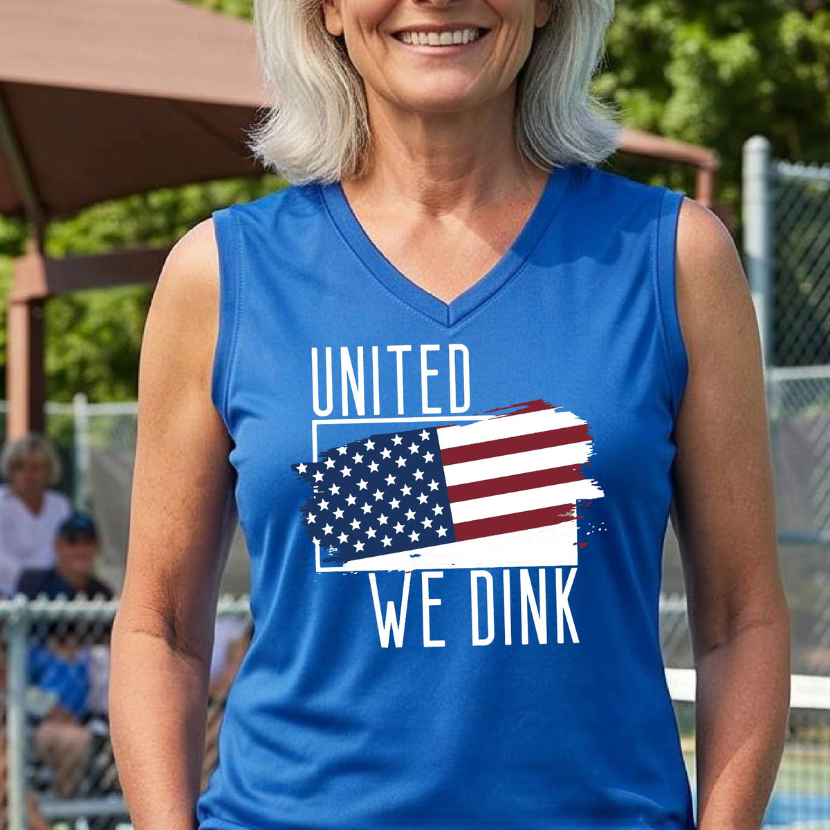 A woman on a pickleball court wearing a royal blue sleeveless t-shirt with a pickleball themed red, white and blue graphic of a patriotic flag with white words “UNITED WE DINK” above and below the flag, on the front of the shirt. 