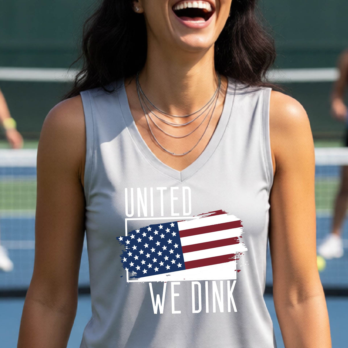 A woman on a pickleball court wearing a silver gray sleeveless t-shirt with a pickleball themed red, white and blue graphic of a patriotic flag with white words “UNITED WE DINK” above and below the flag, on the front of the shirt. 