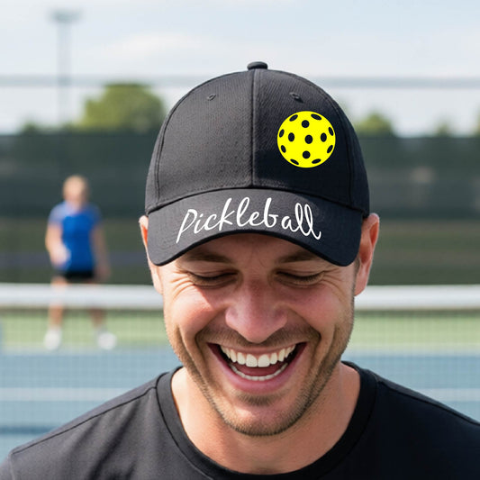 A close-up of a black hat with a graphic of the word “Pickleball” in a white script across the brim of the visor, shown worn in front of a pickleball court in the background. 