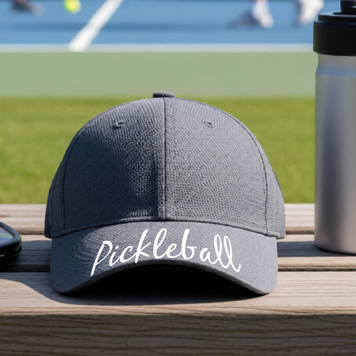 A close-up of a graphite gray hat with a graphic of the word “Pickleball” in a white script across the brim of the visor, shown worn in front of a pickleball court in the background. 