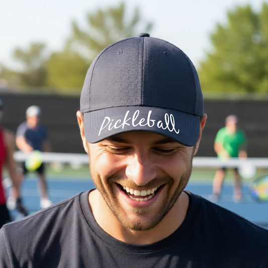 A close-up of a graphite gray hat with a graphic of the word “Pickleball” in a white script across the brim of the visor, shown worn in front of a pickleball court in the background. 