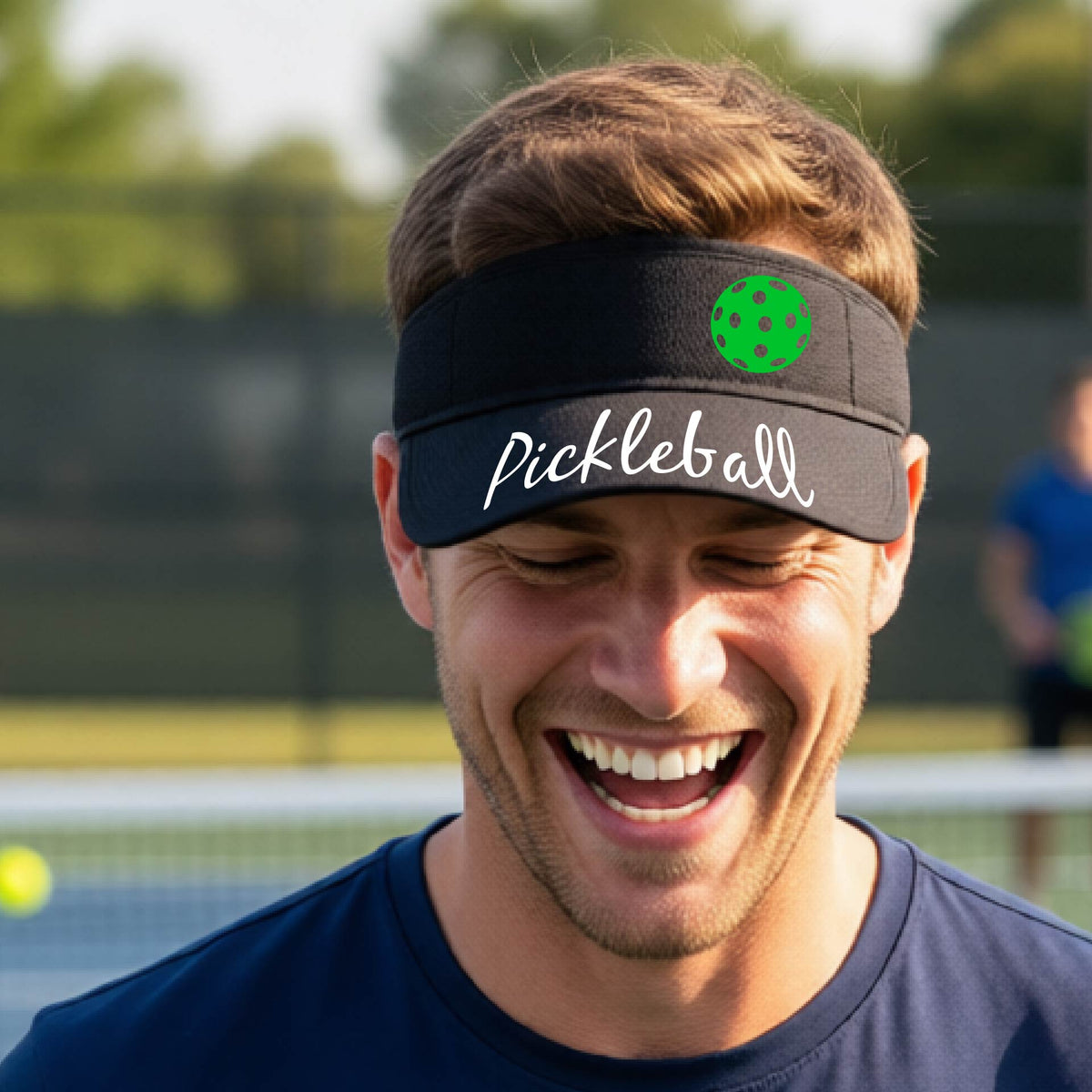 A close-up of a black visor with a graphic of the word “Pickleball” in a white script across the brim of the visor, including a colorful pickleball shown worn in front of a pickleball court in the background. 