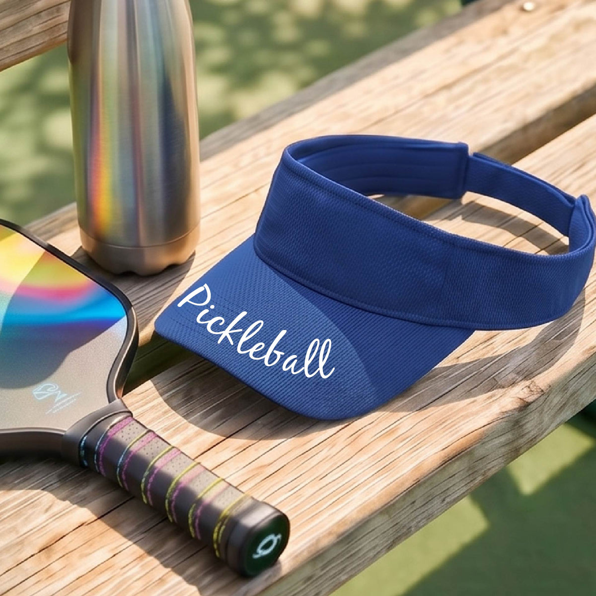 A close-up of a royal blue visor with a graphic of the word “Pickleball” in a white script across the brim of the visor, shown worn in front of a pickleball court in the background. 