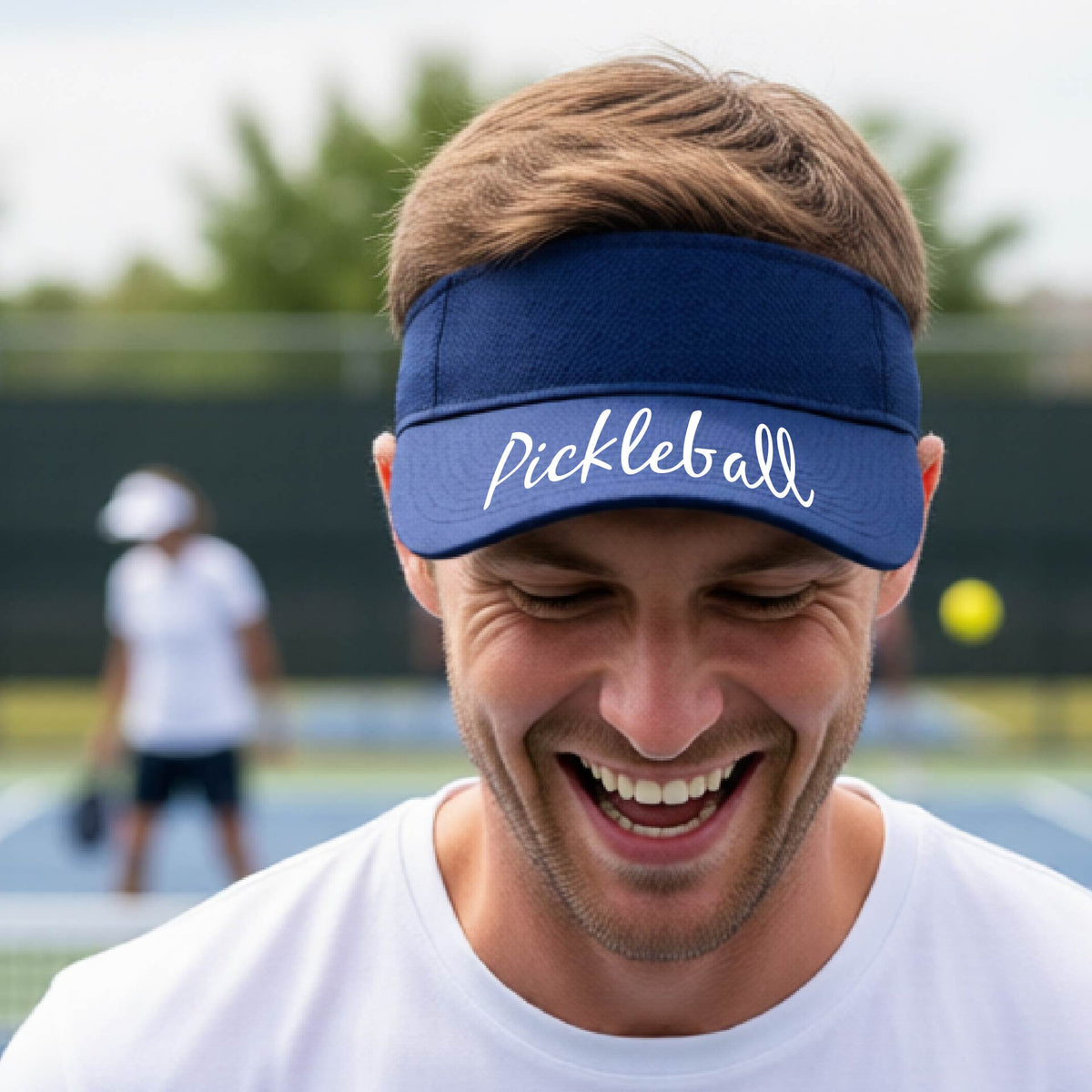 A close-up of a royal blue visor with a graphic of the word “Pickleball” in a white script across the brim of the visor, shown worn in front of a pickleball court in the background. 