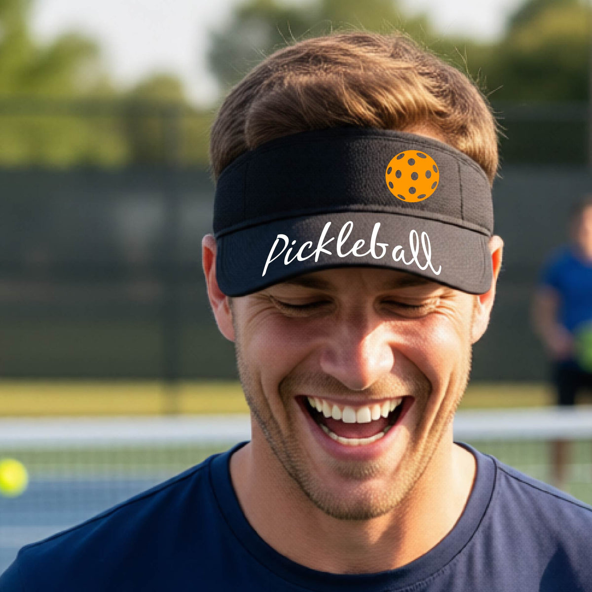 A close-up of a black visor with a graphic of the word “Pickleball” in a white script across the brim of the visor, including a colorful pickleball shown worn in front of a pickleball court in the background. 