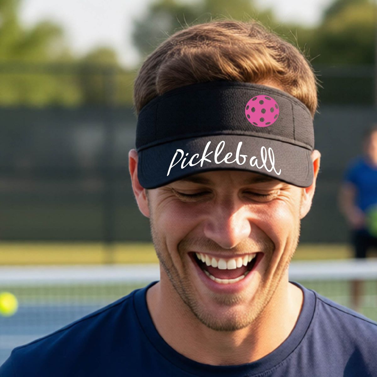 A close-up of a black visor with a graphic of the word “Pickleball” in a white script across the brim of the visor, including a colorful pickleball shown worn in front of a pickleball court in the background. 