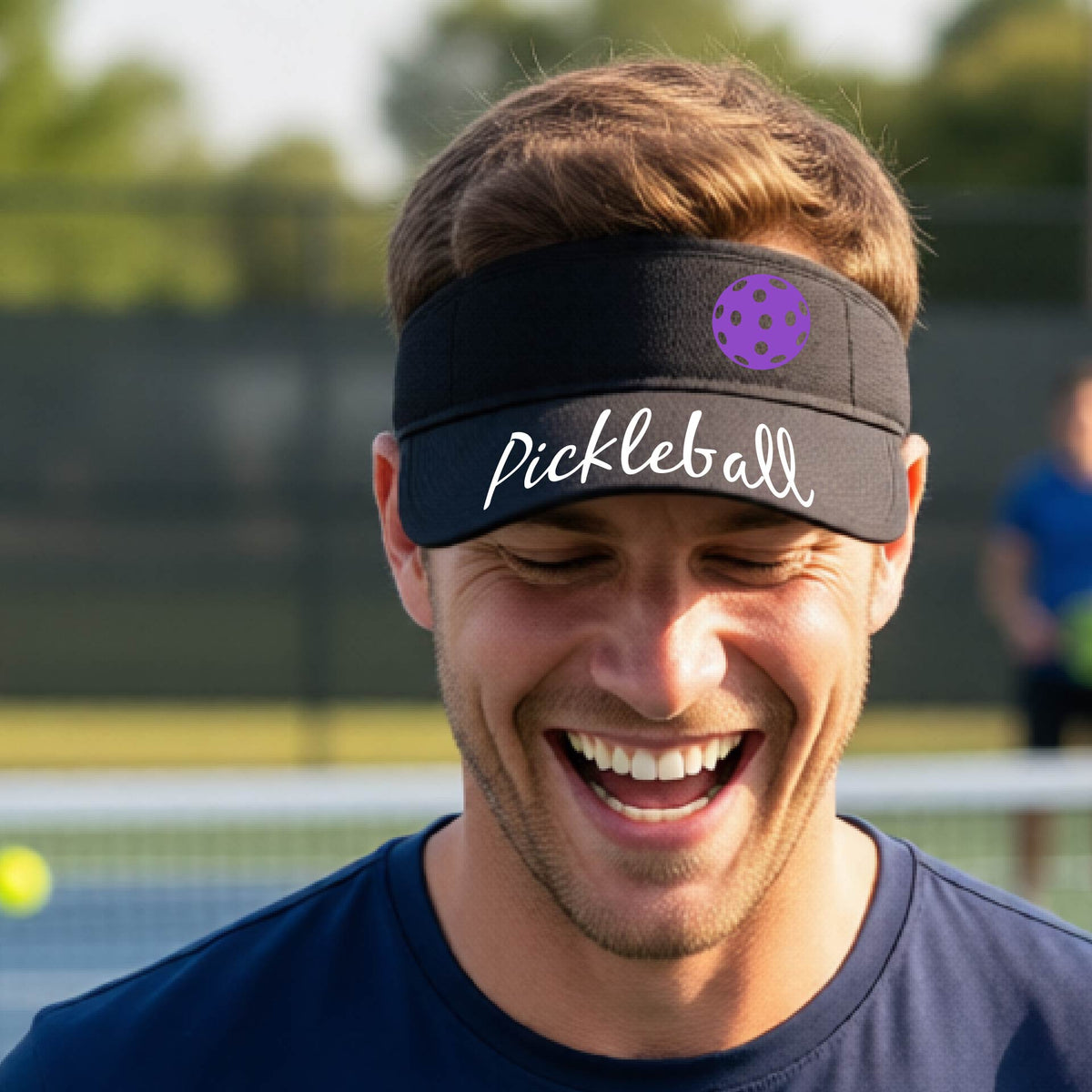 A close-up of a black visor with a graphic of the word “Pickleball” in a white script across the brim of the visor, including a colorful pickleball shown worn in front of a pickleball court in the background. 