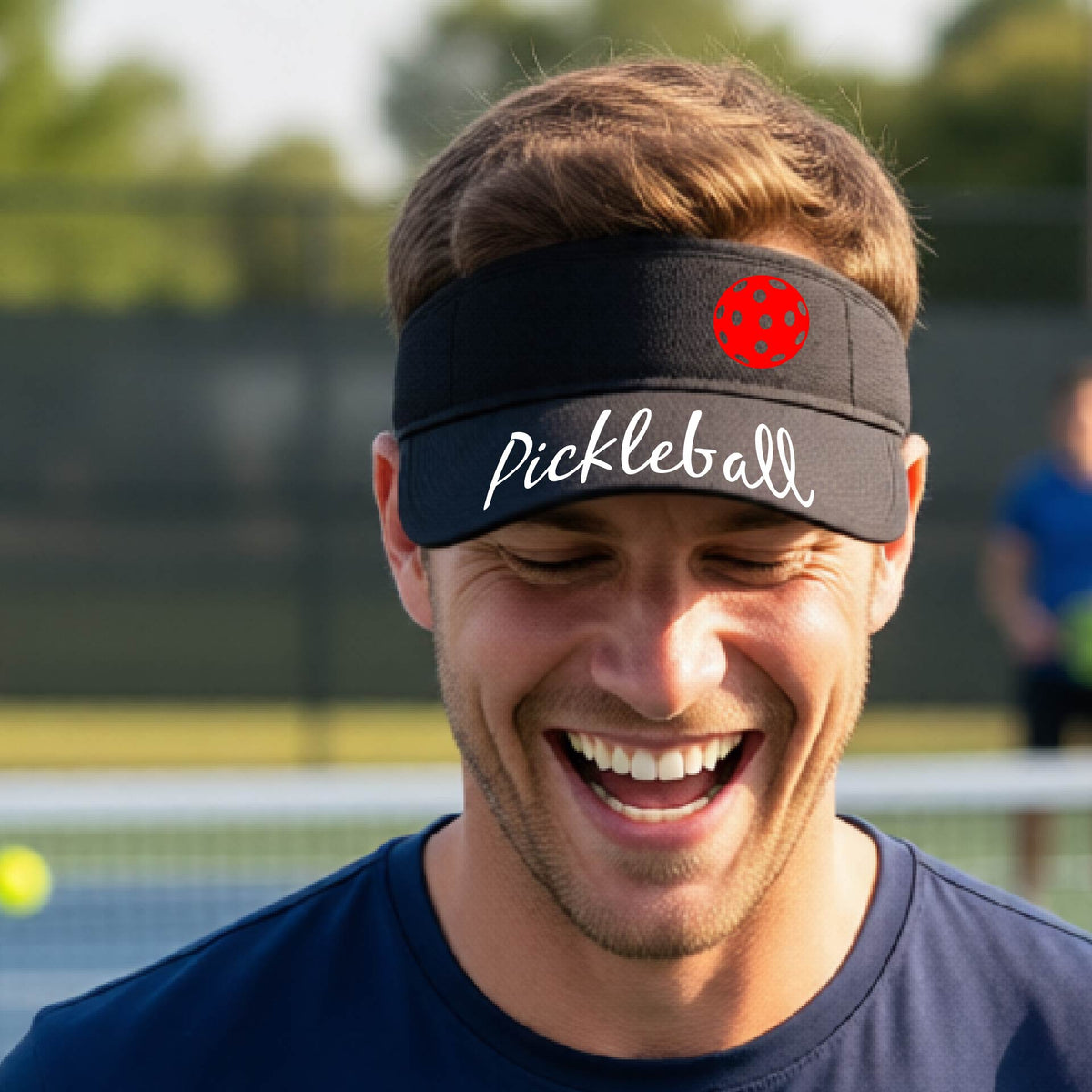 A close-up of a black visor with a graphic of the word “Pickleball” in a white script across the brim of the visor, including a colorful pickleball shown worn in front of a pickleball court in the background. 