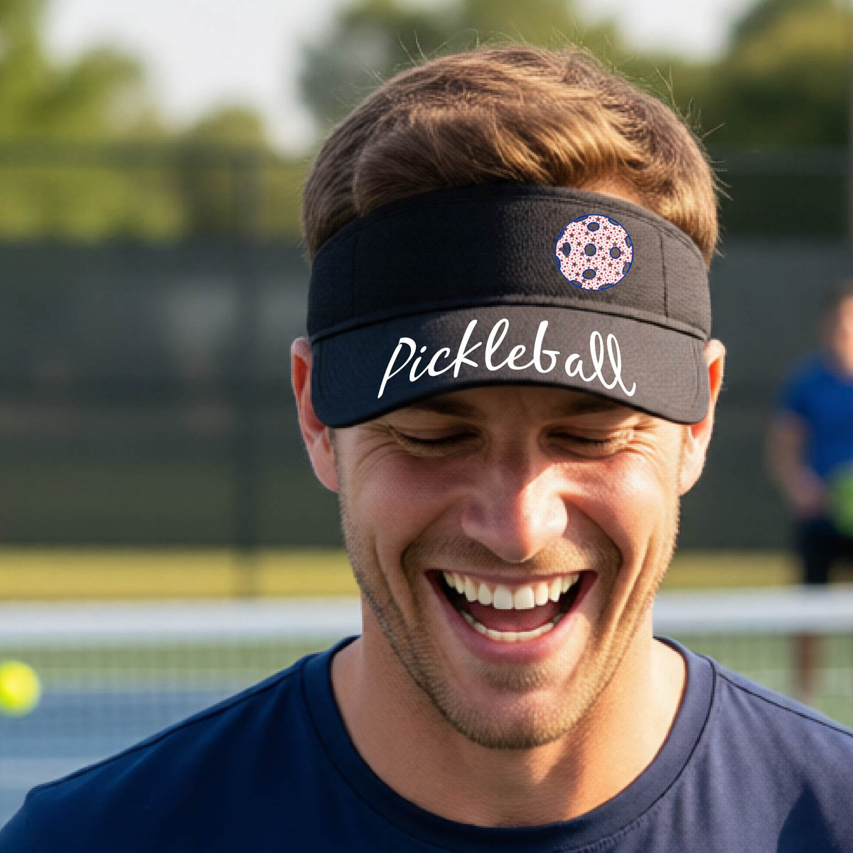 A close-up of a black visor with a graphic of the word “Pickleball” in a white script across the brim of the visor, including a colorful pickleball shown worn in front of a pickleball court in the background. 