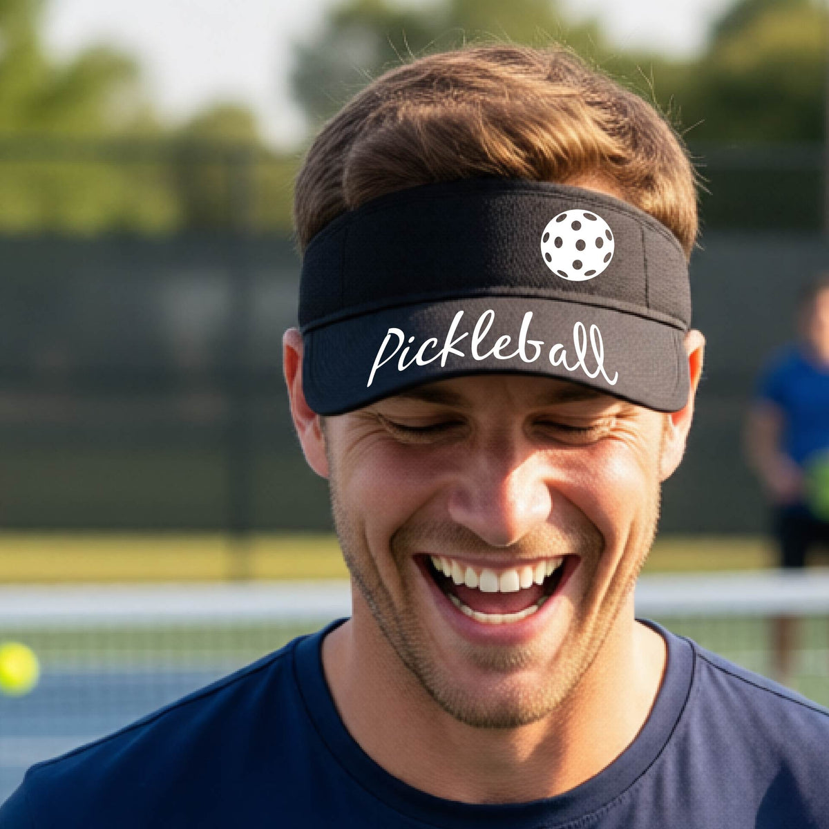 A close-up of a black visor with a graphic of the word “Pickleball” in a white script across the brim of the visor, including a colorful pickleball shown worn in front of a pickleball court in the background. 