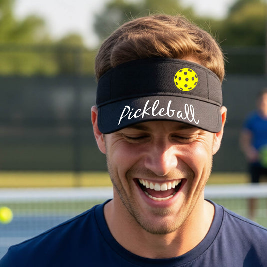 A close-up of a black visor with a graphic of the word “Pickleball” in a white script across the brim of the visor, including a colorful pickleball shown worn in front of a pickleball court in the background. 