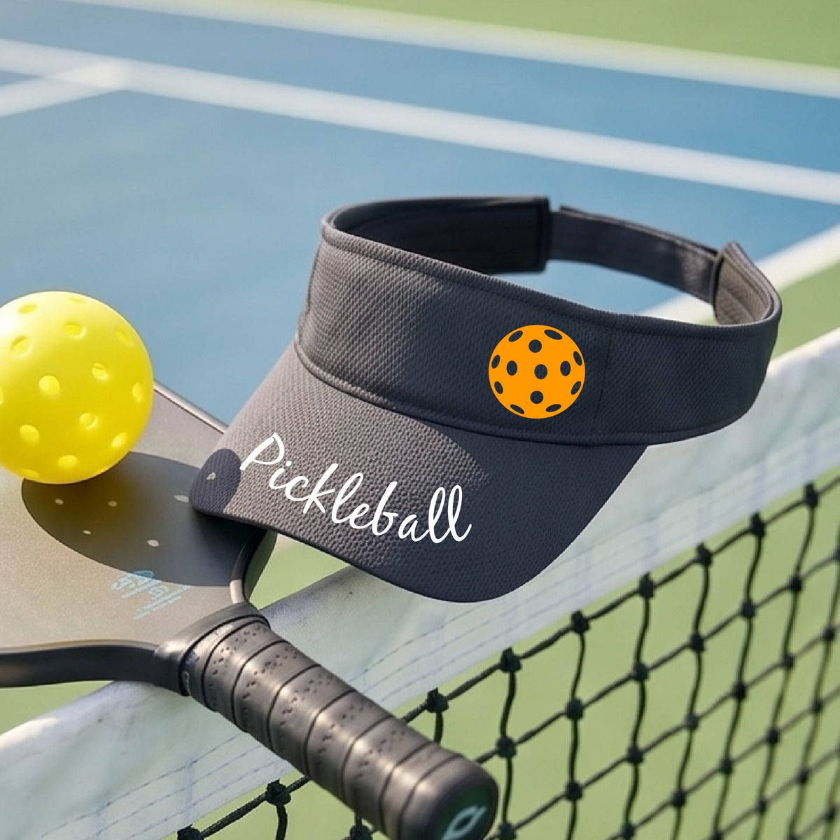 A close-up of a graphite gray visor with a graphic of the word “Pickleball” in a white script across the brim of the visor, including a colorful pickleball shown worn in front of a pickleball court in the background. 