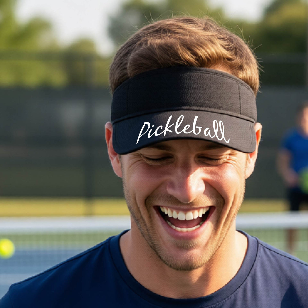 A close-up of a black visor with a graphic of the word “Pickleball” in a white script across the brim of the visor, shown worn in front of a pickleball court in the background. 