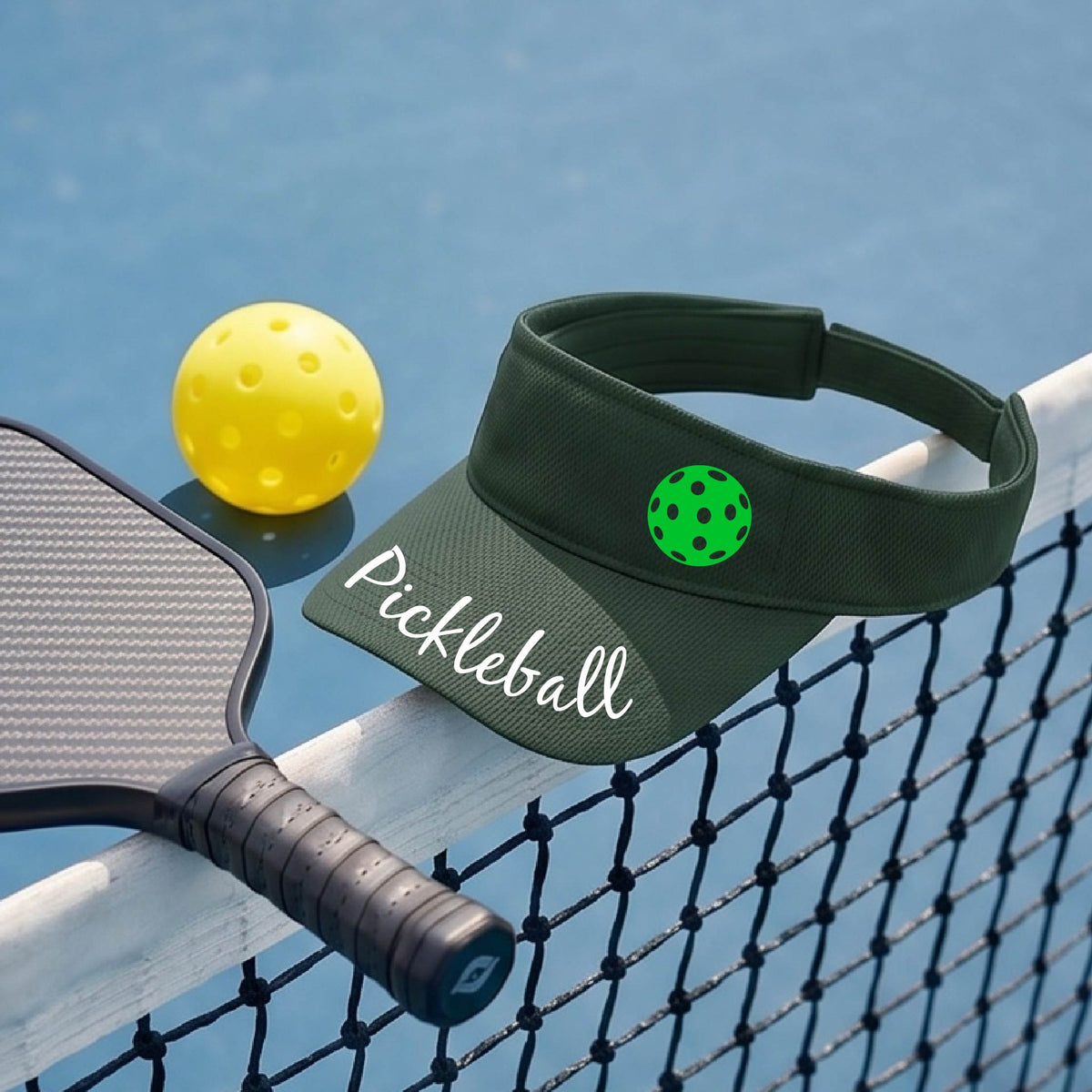 A close-up of a green visor with a graphic of the word “Pickleball” in a white script across the brim of the visor, including a colorful pickleball shown worn in front of a pickleball court in the background. 